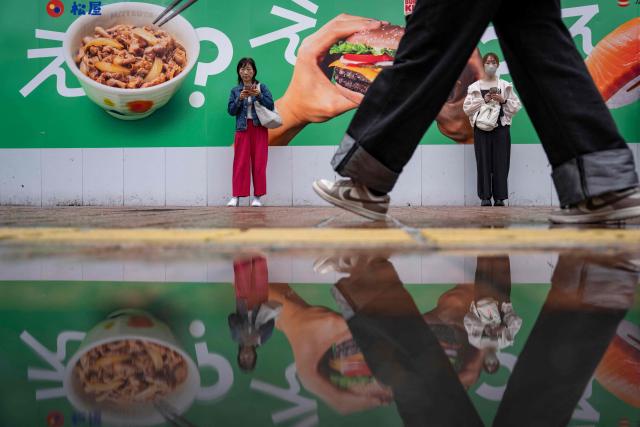 People stand beside a food delivery service advertisement as their reflection shimmers in a puddle in Tokyo on April 27, 2026. (Photo by Yuichi YAMAZAKI / AFP)