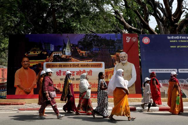 Women walk past a hoarding featuring India's Prime Minister Narendra Modi and chief minister of Uttar Pradesh state Yogi Adityanath, as they arrive to attend a public meeting for women addressed by Modi in Varanasi on April 28, 2026. (Photo by Niharika KULKARNI / AFP)