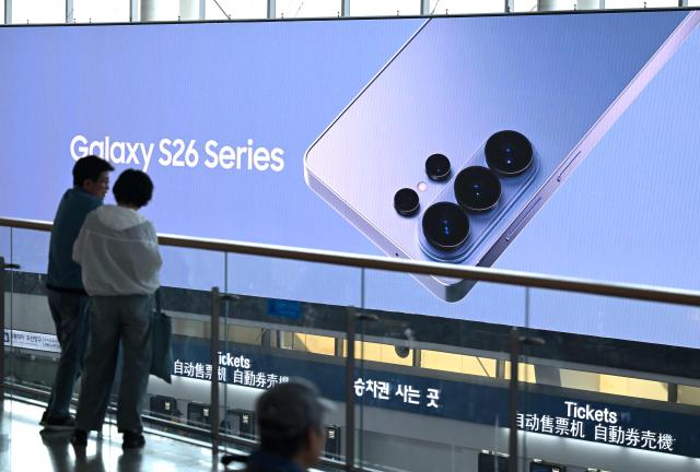 A couple stands in front of a large electronic screen advertising the Samsung Galaxy S26 smartphone at a train station in Seoul on April 30, 2026. South Korean tech giant Samsung Electronics posted a record quarterly profit on April 30, driven by strong sales of chips crucial for artificial intelligence. (Photo by Jung Yeon-je / AFP)