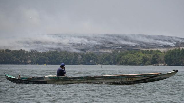A boatman covers his face as he sails past grey smoke billowing from a burning landfill in the nearby Navotas City as seen from Obando town, Bulacan province, on April 30, 2026. The fire at Metro Manila's Navotas landfill is largely invisible to the naked eye, combusting as deep as 15 metres (50 feet) below the surface and releasing a toxic brew of methane as well as carbon dioxide. (Photo by Ted ALJIBE / AFP) / To go with "PHILIPPINES-FIRE-WASTE-ENVIRONMENT-POLLUTION, REPORTAGE" BY PAM CASTRO