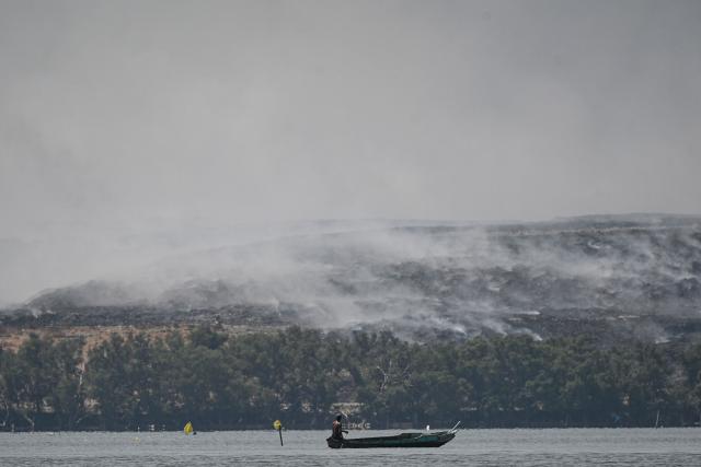A fisherman sails past grey smoke billowing from a burning landfill in the nearby Navotas City as seen from Obando town, Bulacan province, on April 30, 2026. The fire at Metro Manila's Navotas landfill is largely invisible to the naked eye, combusting as deep as 15 metres (50 feet) below the surface and releasing a toxic brew of methane as well as carbon dioxide. (Photo by Ted ALJIBE / AFP) / To go with "PHILIPPINES-FIRE-WASTE-ENVIRONMENT-POLLUTION, REPORTAGE" BY PAM CASTRO