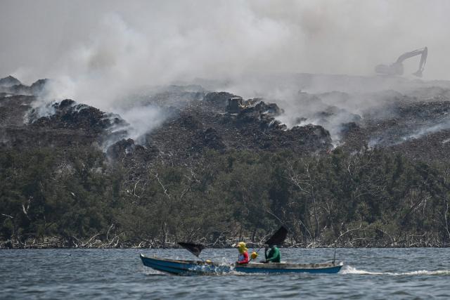Fishermen sail past grey smoke billowing from a burning landfill in the nearby Navotas City as seen from Obando town, Bulacan province, on April 30, 2026. The fire at Metro Manila's Navotas landfill is largely invisible to the naked eye, combusting as deep as 15 metres (50 feet) below the surface and releasing a toxic brew of methane as well as carbon dioxide. (Photo by Ted ALJIBE / AFP) / To go with "PHILIPPINES-FIRE-WASTE-ENVIRONMENT-POLLUTION, REPORTAGE" BY PAM CASTRO