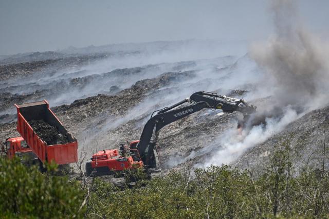 Heavy equipment work on a burning landfill in the nearby Navotas City, as seen from a boat along the coast of Obando town, Bulacan province, on April 30, 2026. The fire at Metro Manila's Navotas landfill is largely invisible to the naked eye, combusting as deep as 15 metres (50 feet) below the surface and releasing a toxic brew of methane as well as carbon dioxide. (Photo by Ted ALJIBE / AFP) / To go with "PHILIPPINES-FIRE-WASTE-ENVIRONMENT-POLLUTION, REPORTAGE" BY PAM CASTRO