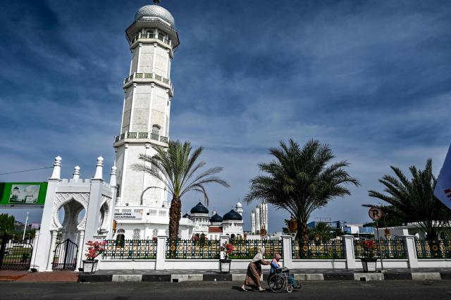 A woman is pushed in a wheelchair past a mosque in Banda Aceh on May 1, 2026. (Photo by CHAIDEER MAHYUDDIN / AFP)