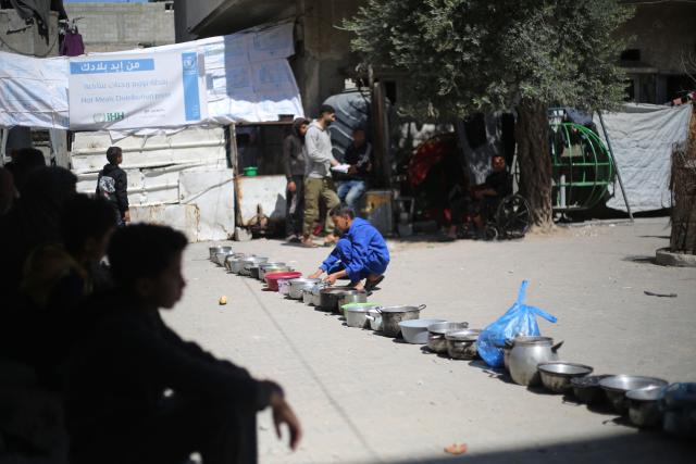 Displaced Palestinians sit waiting near a row of empty food containers placed near the rubble of buildings destroyed by the Israeli military, as families wait to collect food from a charity kitchen in the Bureij refugee camp, in the central Gaza Strip on May 1, 2026. The war on the Gaza Strip was sparked following the militant Palestinian group Hamas's October 7, 2023 attack on Israel, which resulted in the deaths of 1,221 people. Israel's retaliatory assault on Gaza has killed at least 72,600 people, according to figures from the health ministry that the UN considers reliable. with th  majority of Gaza's 2.4 million people being displaced. (Photo by Eyad Baba / AFP)