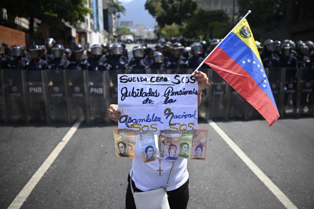 TOPSHOT - A woman holds a sign reading "Zero salary, SOS retirees and pensioners of the National Assembly" as she carries a Venezuelan flag and bolivar banknotes in front of members of the Bolivarian National Police Corps (CPNB) during a May Day rally demanding better working conditions and pensions, marking International Workers Day, in Caracas, on May 1, 2026. (Photo by Federico PARRA / AFP)