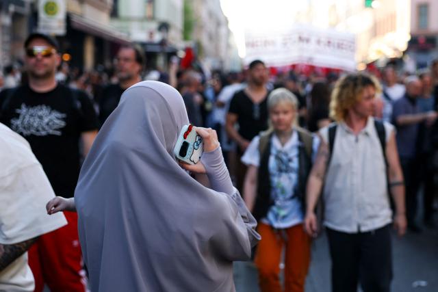 A woman wearing a headscarf and holding a child is seen holding a mobile phone as people walk in the streets during the annual leftist May Day demonstration in Berlin's Kreuzberg district on May 1, 2026. (Photo by Odd ANDERSEN / AFP)