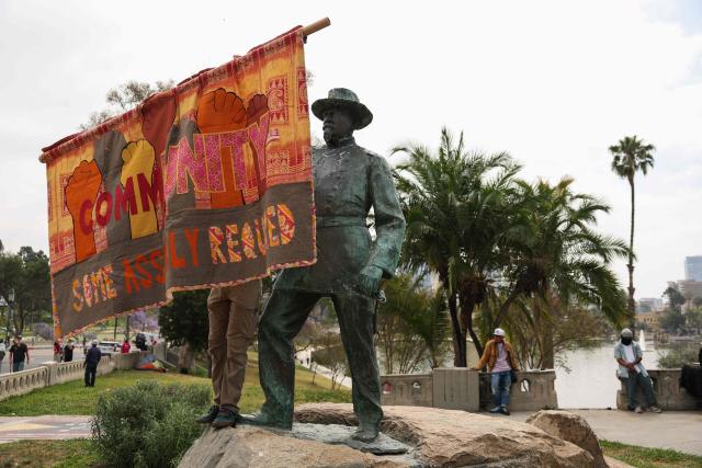 A demontrator holds a banner by the statue of General Harrison Gray Otis in MacArthur Park as people march during a May Day rally marking International Workers' Day in Los Angeles, California, on May 1, 2026. (Photo by Patrick T. Fallon / AFP)