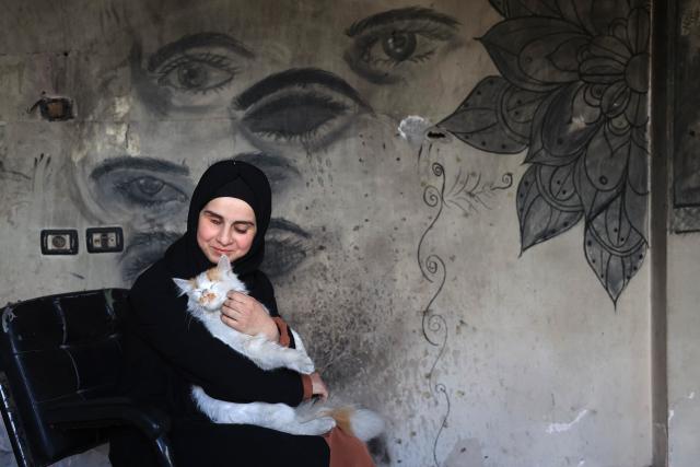 Areej al-Saafin, who lost her eye in an Israeli military strike, holds her pet cat as she sits in front of her charcoal drawings that decorate the wall of her home in the Bureij Palestinian refugee camp in the central Gaza Strip on May 2, 2026. The war on the Gaza Strip was sparked following the militant Palestinian group Hamas's October 7, 2023 attack on Israel, which resulted in the deaths of 1,221 people. Israel's retaliatory assault on Gaza has killed at least 72,600 people, according to figures from the health ministry that the UN considers reliable, with the majority of Gaza's 2.4 million people being displaced. (Photo by Eyad Baba / AFP)