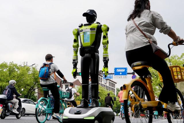 People cycle past a humanoid robot police officer in Hangzhou, in China’s eastern Zhejiang province on May 3, 2026 on the third day of a five-day national May Day holiday. The tech hub's traffic police deployed 15 humanoid robots in total to bolster their ranks on May 1. (Photo by Agatha CANTRILL / AFP)