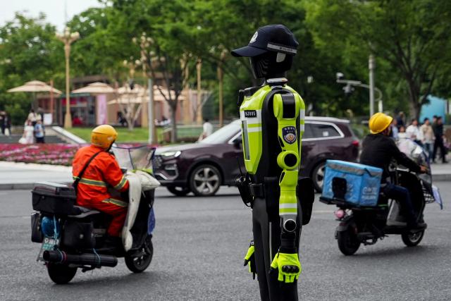 People ride past a humanoid robot police officer in Hangzhou, in China’s eastern Zhejiang province on May 3, 2026 on the third day of a five-day national May Day holiday. The tech hub's traffic police deployed 15 humanoid robots in total to bolster their ranks on May 1. (Photo by Agatha CANTRILL / AFP)