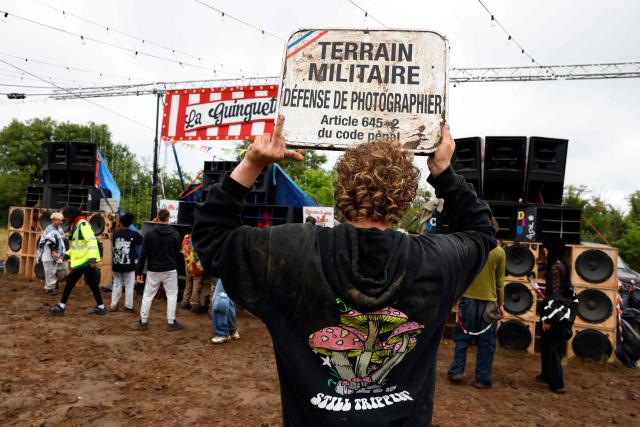 A partygoer gestures as he holds a placard reading "military site; photography prohibited" at the end of a "teknival" rave party at a military site in Cornusse, near Bourges, central France on May 4, 2026. Between 17,000 and 40,000 people participate in the free party which has been taking place, without major incident, since May 1, 2026. The gathering comes as the French parliament is seeking to tighten legislation against unauthorised rave parties, introducing prison sentences for organisers and fines for attendees. (Photo by Kenzo TRIBOUILLARD / AFP)