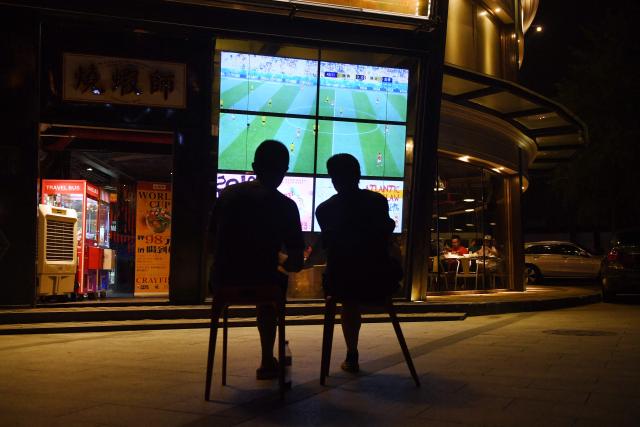 (FILES) This photo taken on July 3, 2018 shows two football fans watching the 2018 Russia World Cup football match between Sweden and Switzerland on a TV screen outside a restaurant in Beijing. Swathes of football-mad Asia from China to India, Malaysia and Thailand, are yet to announce broadcast rights for the biggest-ever World Cup with kick-off barely one month away. (Photo by GREG BAKER / AFP)