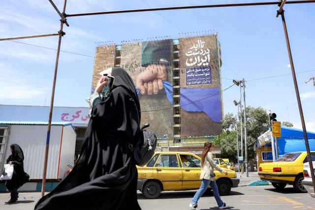 A woman adjusts her headdress as she walks near a billboard on the facade of a building depicting the Strait of Hormuz with a caption in Persian reading "Forever in Iran’s Hand", at Vanak Square in Tehran on May 6, 2026. The United States and Pakistan, a key mediator in the Middle East war, expressed new hope of talks leading to a peace deal with Iran on May 6, after President Donald Trump halted a plan to guide ships through the Strait of Hormuz. (Photo by AFP) / 