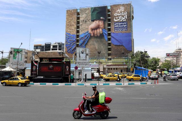 A motorist rides a scooter along a street near a billboard on the facade of a building depicting the Strait of Hormuz with a caption in Persian reading "Forever in Iran’s Hand", at Vanak Square in Tehran on May 6, 2026. The United States and Pakistan, a key mediator in the Middle East war, expressed new hope of talks leading to a peace deal with Iran on May 6, after President Donald Trump halted a plan to guide ships through the Strait of Hormuz. (Photo by AFP) / 