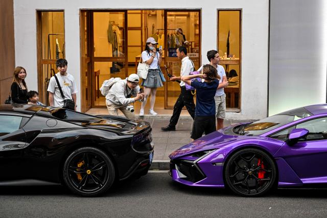 A man takes photographs of a young man next to Ferrari and Lamborghini supercars on a street in Shanghai on May 6, 2026. (Photo by Hector RETAMAL / AFP)
