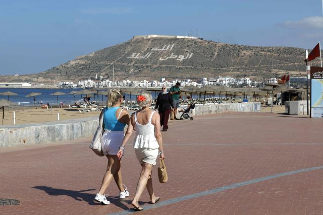 The words ‘Allah, Country and the King’ is seen on the hillside overlooking the Agadir Corniche that runs along the Atlantic Ocean, as two women walk along the promenade in the city of Agadir on May 7, 2026. This promenade is very popular with locals, visitors, and tourists, especially for walking, sports, and leisure activities. (Photo by Abdel Majid BZIOUAT / AFP)