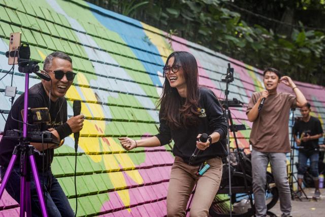 Online buskers use a colourful wall as a backdrop as they perform on a sidewalk to attract viewers for digital donations during their livestreams on social media sites in Bandung, West Java, on May 8, 2026. The singers performing on social media video streaming sites earn on average 4-10 USD from digital payments over three hours of performing. (Photo by TImur Matahari / AFP)