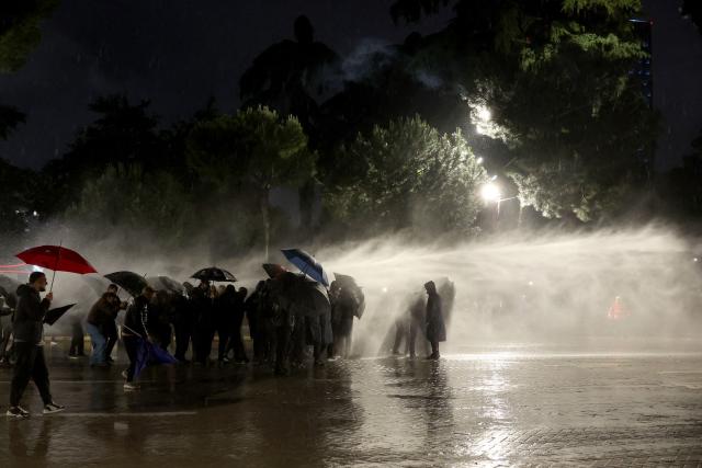 Demonstrators protect themselves from water sprayed by a riot police water cannon truck during an anti-government protest in Tirana on May 8, 2026. (Photo by Adnan Beci / AFP)