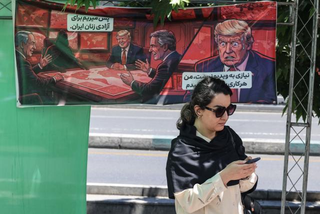 An Iranian woman looks at her mobile phone as she walks past a banner depicting a cartoon of the US president which reads, 'So we go for the freedom of Iranian women' (L) and 'There is no need for a video, I will do what ever you say' (R),  in the capital Tehran on May 9, 2026. Iran questioned the seriousness of American diplomacy on May 9, in the wake of renewed naval clashes in the Gulf, while keeping Washington waiting for a response to its latest negotiating position. On February 28, 2026, Israel and the United States launched strikes on Iran, killing its supreme leader and triggering a war that spread across the Middle East. (Photo by ATTA KENARE / AFP) / 
