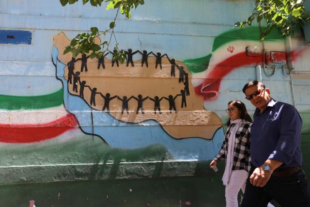An Iranian man amd woman walk past a mural of the map of Iran, with its citizens holding hands, painted on a wall in the capital Tehran on May 9, 2026. Iran questioned the seriousness of American diplomacy on May 9, in the wake of renewed naval clashes in the Gulf, while keeping Washington waiting for a response to its latest negotiating position. On February 28, 2026, Israel and the United States launched strikes on Iran, killing its supreme leader and triggering a war that spread across the Middle East. (Photo by ATTA KENARE / AFP) / 