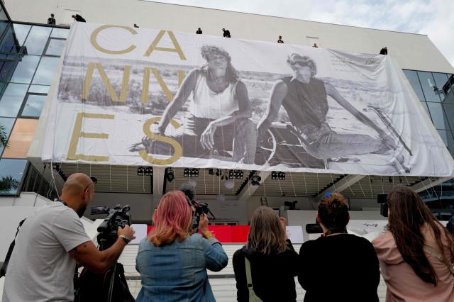 Workers hang the official poster of the 78th Cannes Film Festival on the facade of the Palais des Festivals as journalists use cameras to catch footage in Cannes, south-eastern France on May 10, 2026. The Cannes Film festival will take place from May 12 to May 23, 2026. (Photo by Valery HACHE / AFP)
