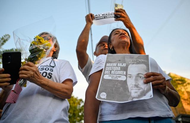 A woman holds a sign depicting late Venezuelan political prisoner Victor Hugo Quero Navas during Mother's Day outside El Rodeo I prison in Guatire, Miranda state, Venezuela on May 10, 2026. Venezuelan authorities exhumed the body of Victor Hugo Quero Navas, a political prisoner who died in July 2025 but whose death was not officially acknowledged until May 7, 2026, prompting criticism from civil society and the opposition, an AFP journalist reported. (Photo by Juan BARRETO / AFP)