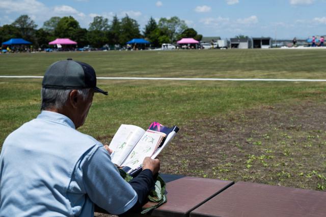 In this photo taken on May 9, 2026, a spectator reads a cricket rule book as he watches the game during the ICC T20 World Cup East Asia-Pacific Qualifier match between Japan and Vanuatu at the Nisshin Cricket Ground at Korogi Sports Park in Nisshin, Aichi Prefecture. Curious spectators clutching rulebooks watch as a batsman smacks a six into the sand and weeds surrounding Japan's brand-new cricket ground, four months before it hosts matches at the Asian Games. (Photo by Andrew CABALLERO-REYNOLDS / AFP) / To go with 'ASIAD-2026-CRICKET-JPN,FOCUS' by Andrew MCKIRDY