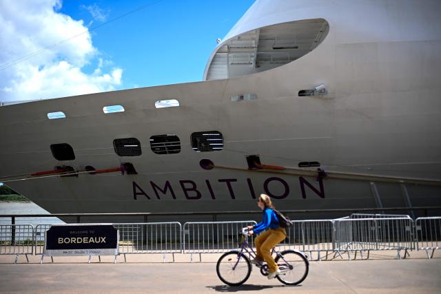 A woman rides a bicycle past the Bahamas-registered cruise ship Ambition after passengers were confined following the outbreak of a gastrointestinal illness onboard, at the Bordeaux port in Bordeaux, southwestern France on May 13, 2026. French authorities on May 13 confined more than 1,700 passengers and crew on a British cruise ship docked in Bordeaux after an elderly passenger died, said officials, who played down any links to the hantavirus scare. (Photo by Christophe ARCHAMBAULT / AFP)
