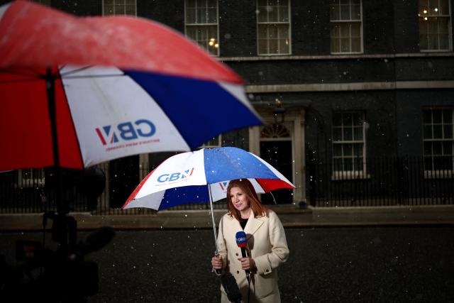 A member of the media shelters under an umbrella at Downing Street in central London on May 13, 2026. Britain's Prime Minister Keir Starmer, fighting to face down a revolt within his ruling Labour party, has promised his government will be "better" and bolder to assuage disgruntled voters impatient for change. (Photo by Henry Nicholls / AFP)