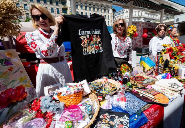Ukrainian women, dressed in traditional costumes, hold up a T-shirt with the ESC competitors, during the ESC side event 'Eurovision Market Contest' at Vienna's Naschmarkt, Austria, where several food and souvenir stalls from different ESC participating countries take part, on May 13, 2026. (Photo by Joe KLAMAR / AFP)