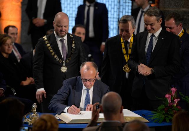 German Chancellor Friedrich Merz (C) signs the golden book as Greek Prime Minister Kyriakos Mitsotakis (R), the former President of the European Central Bank (ECB) and former Italian Prime Minister Mario Draghi (2nd R) and Aachen's mayor Michael Ziemons (L) look on after Draghi was awarded the 2026 International Charlemagne Prize of Aachen, on May 14, 2026 in Aachen, western Germany. (Photo by Sascha Schuermann / AFP)