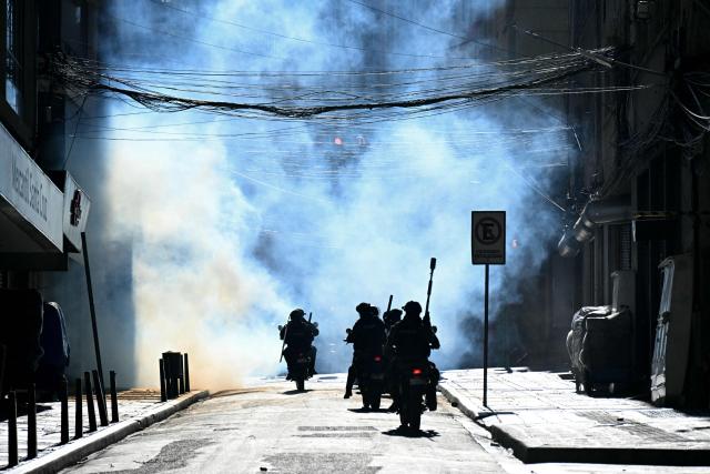 Riot police on motorcycles chase miners through tear gas during a march to demand the resignation of Bolivian President Rodrigo Paz, in La Paz, on May 14, 2026. For the past two weeks, workers, farmers, school teachers, Indigenous groups and transport workers have been pressuring the government. Their demands range from wage increases and economic stabilization to opposition to the privatization of public companies. (Photo by AIZAR RALDES / AFP)