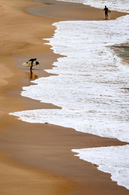 A surfer crouches as he carries his board after walking out of the surf at Bondi Beach on a cold autumn day in Sydney on May 18, 2026. (Photo by DAVID GRAY / AFP)
