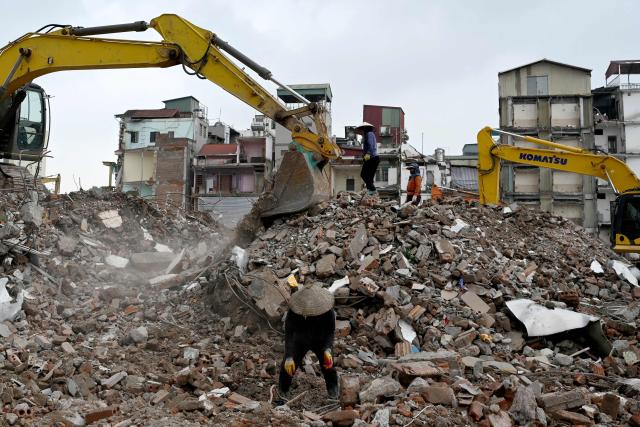Workers (C) collect scrap metal from the rubble of residential buildings as excavators are seen at a demolition site to make way for an expanded road network in Hanoi on May 18, 2026. (Photo by Nhac NGUYEN / AFP)