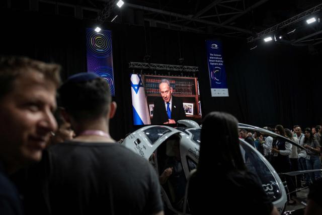 The of Israeli Prime Minister Benjamin Netanyahu is seen on a large screen as visitors inspect a 'flying pod' at the Smart Mobility Summit 2026, in Tel Aviv on May 18, 2026. (Photo by ilia YEFIMOVICH / AFP)