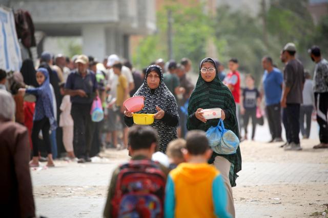 Displaced Palestinian women carry pots of food collected from a charity kitchen to their family shelter as they walk along a former road destroyed by the Israeli military in the Bureij refugee camp in the central Gaza Strip on May 18, 2026. The war has displaced nearly Gaza's entire population and reduced swathes of the Palestinian territory to rubble despite a ceasefire in October 2025, the enclave remains gripped by daily violence as Israeli strikes continue, with both the military and Hamas accusing one another of violating the truce. Hundreds of thousands of people are still living in tents and conditions remain dire despite a ceasefire that took effect in October last year. (Photo by Eyad Baba / AFP)