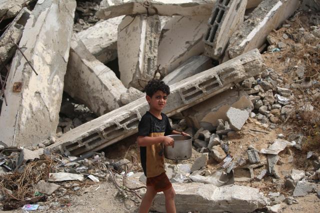 A displaced Palestinian child carries a pot of food distributed by a charity kitchen, past buildings destroyed by the Israeli military, in the Bureij refugee camp, in the central Gaza Strip on May 18, 2026. The war has displaced nearly Gaza's entire population and reduced swathes of the Palestinian territory to rubble despite a ceasefire in October 2025, the enclave remains gripped by daily violence as Israeli strikes continue, with both the military and Hamas accusing one another of violating the truce. Hundreds of thousands of people are still living in tents and conditions remain dire despite a ceasefire that took effect in October last year. (Photo by Eyad Baba / AFP)