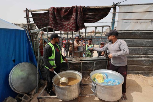 Two volunteers at a charity kitchen stand near huge pots as they dish out food to waiting displaced Palestinian families in the Bureij refugee camp, in the central Gaza Strip on May 18, 2026. The war has displaced nearly Gaza's entire population and reduced swathes of the Palestinian territory to rubble despite a ceasefire in October 2025, the enclave remains gripped by daily violence as Israeli strikes continue, with both the military and Hamas accusing one another of violating the truce. Hundreds of thousands of people are still living in tents and conditions remain dire despite a ceasefire that took effect in October last year. (Photo by Eyad Baba / AFP)