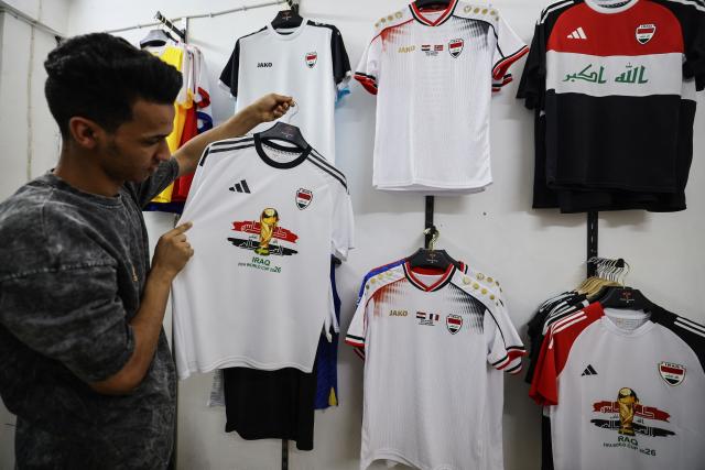A man displays a football jersey bearing the FIFA World Cup Trophy and the Iraqi national flag at his sportswear shop in Baghdad on May 18, 2026, ahead of the 2026 FIFA World Cup, for which the country qualified. The "Lions of Mesopotamia" secured the country's first World Cup appearance since the 1986 competition in Mexico with a 2-1 win over Bolivia on March 31, 2026. (Photo by AHMAD AL-RUBAYE / AFP)