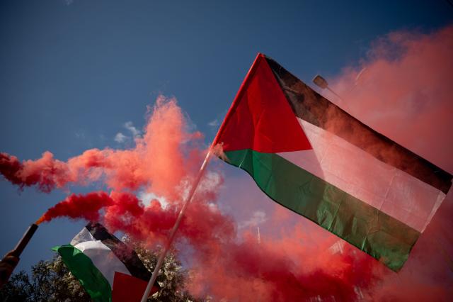 A Palestinian flag is pictured against a background of smoke grenades during a gathering against the interception of the Global Sumud Flotilla by the Israeli Navy off the coast of Cyprus outside the Ministry of Foreign Affairs in Athens on May 18, 2026. Israeli forces intercepted a Gaza-bound aid flotilla on May 18, 2026 after it sailed from Turkey the previous week, with Prime Minister Benjamin Netanyahu denouncing the mission as a "malicious scheme" intended to support Hamas, as the Global Sumud Flotilla vessels mark the latest in a series of attempts by activists to breach Israel's blockade of the Palestinian territory, with the last convoy intercepted by Israeli forces the previous month. (Photo by Aggelos NAKKAS / AFP)