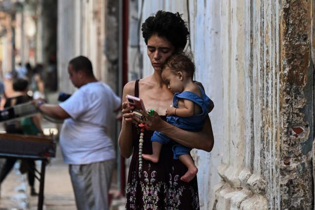 A woman carries her baby while looking at her mobile phone in Havana on May 20, 2026. (Photo by YAMIL LAGE / AFP)