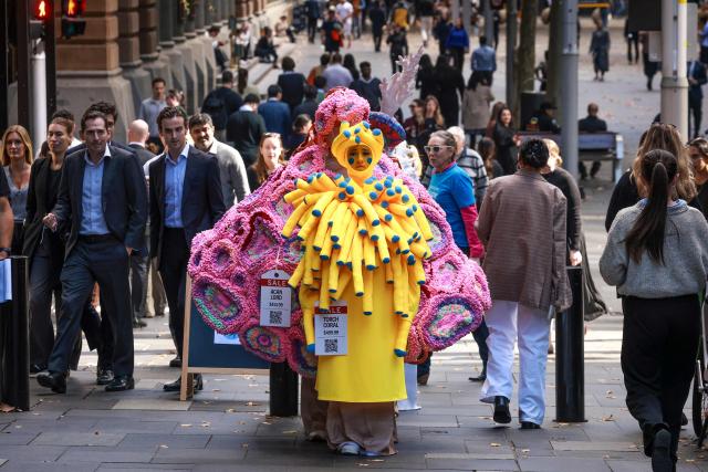 Performers wear coral reef-inspired costumes on a footpath next to pedestrians during a show to raise awareness of threats to Australia's Great Barrier Reef in Sydney on May 21, 2026. (Photo by DAVID GRAY / AFP)