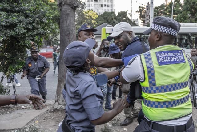 Members of the South African Police Service (SAPS) escort a foreign national resisting to get into a bus from the Diakonia Centre to the Home Affairs offices in Durban on May 21, 2026. The crowd, made up largely of refugees and foreign nationals had been sheltering at the station for up to two days, seeking police protection amid threats and intimidation. Their presence came against the backdrop of sporadic anti-illegal immigration protests that have unsettled parts of South Africa in recent weeks. (Photo by RAJESH JANTILAL / AFP)