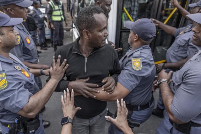 TOPSHOT - Members of the South African Police Service (SAPS) escort a foreign national resisting to get into a bus from the Diakonia Centre to the Home Affairs offices in Durban on May 21, 2026. The crowd, made up largely of refugees and foreign nationals had been sheltering at the station for up to two days, seeking police protection amid threats and intimidation. Their presence came against the backdrop of sporadic anti-illegal immigration protests that have unsettled parts of South Africa in recent weeks. (Photo by RAJESH JANTILAL / AFP)