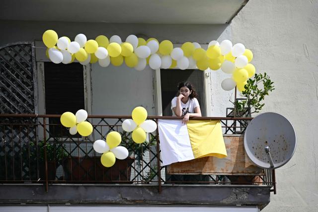 A young girl stands on a balcony overlooking Piazza Nicola Calipari before the arrival of Pope Leo XIV during a pastoral visit to the community of the 'Land of Fires' on May 23, 2026. The Land of Fires (terra dei fuochi) is an area in the Southern Italian region of Campania were, systematically, since the end of the 80s, toxic wastes have been dumped by organized crime (Camorra) causing waste-related environmental crimes. Acerra and the surrounding territory has higher-than-average cancer rates, linked to the dumping, burning, and burying of toxic waste. (Photo by Filippo MONTEFORTE / AFP)