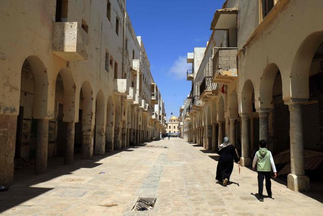 Libyans walk along a bullet scarred street during the reconstruction of the historical centre, partially destroyed during the country's civil war (2014-2017), in the Old City of Benghazi, situated in eastern Libya along the Mediterranean coast, on May 23, 2026. Libya remains plagued by division and instability after years of unrest following the NATO-backed uprising that toppled longtime leader Muammar Gaddafi in 2011. Libya is still divided between the UN-recognised government in the west and its eastern rival in the east, backed by military commander Khalifa Haftar. (Photo by Abdullah DOMA / AFP)
