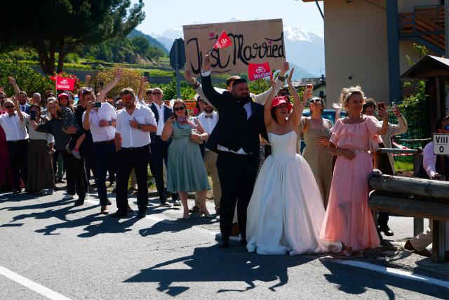 TOPSHOT - Participants of a wedding party holding a placard "Just married" behind a groom and a bride, cheer at riders during a climb in the 14th stage of the Giro d'Italia 2026 - Tour of Italy cycling race between Aosta and Pila (Gressan), Italy, on May 23, 2026. (Photo by Luca Bettini / AFP)