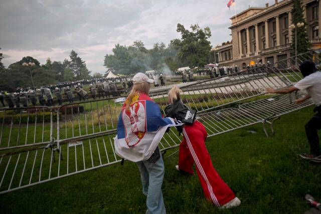 A person wraps in a Serbian flag and two fellow demonstrators push barriers set up by riot police outside Serbia's parliament building during clashes that erupt at the end of an anti-government protest decrying corruption and calling for early elections following the collapse of a railway station canopy in Novi Sad that killed 16 in November 2024, in central Belgrade on May 23, 2026. (Photo by OLIVER BUNIC / AFP)