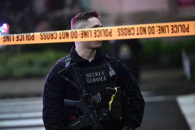 An armed secret service police officer stands behing tape blocking off a crime scene near the White House in Washington, DC, on May 23, 2026. Police and security forces swarmed the area around the White House on the evening of May 23 after reports of shots fired, AFP journalists said. US President Donald Trump was at the White House at the time as he worked to negotiate a deal with Iran. (Photo by Brendan SMIALOWSKI / AFP)